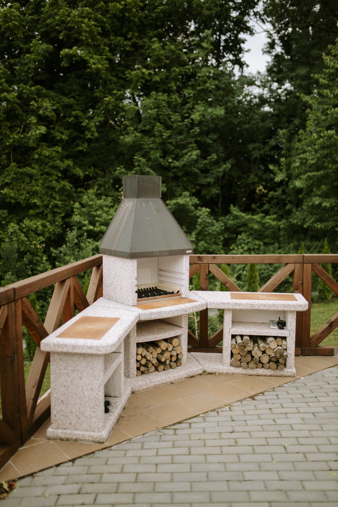 Stone outdoor grill with chimney and built-in benches, surrounded by stacked firewood and green forest backdrop.